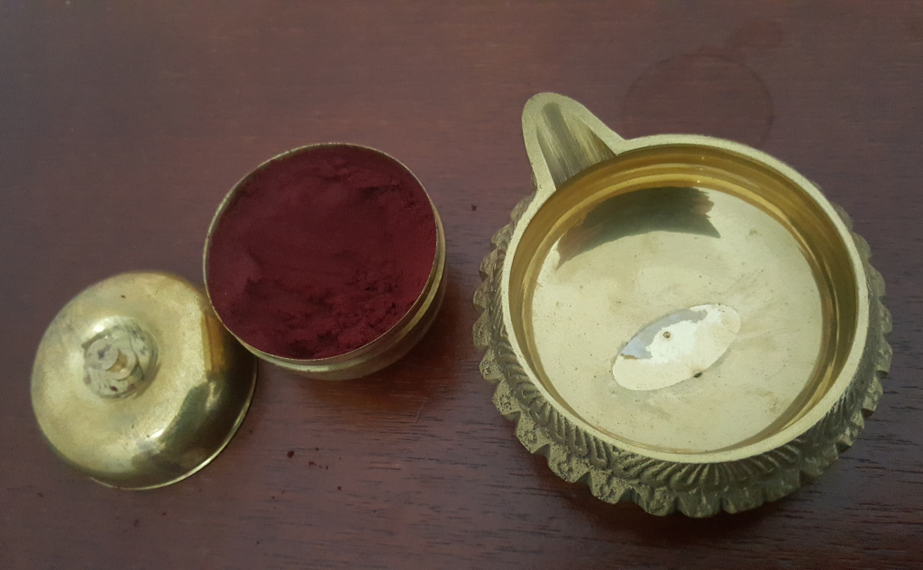 Two brass containers and a dark reddish-brown powder. One container is a small, round, lidded jar with a slightly domed lid. It's filled almost to the brim with a fine, deep crimson powder, which appears smooth and slightly reflective. The powder's consistency looks like finely ground pigment or spice. This jar is positioned slightly off-centre to the left. The lid is separate and resting next to it. The second container is larger, shallow, and more ornate. It resembles a traditional oil lamp or diya, with a spout at one end. The body is decorated with an intricate, raised relief pattern around the rim. It's empty except for a faint, off-white oval mark or stain on the bottom, suggesting possible wear or a previous residue. This container sits off-centre to the right, slightly angled away from the viewer. Both brass pieces appear aged, with subtle variations in tone and highlights indicating the metal's tarnish and polishing. The metallic surfaces have a soft sheen, reflecting a little ambient light.