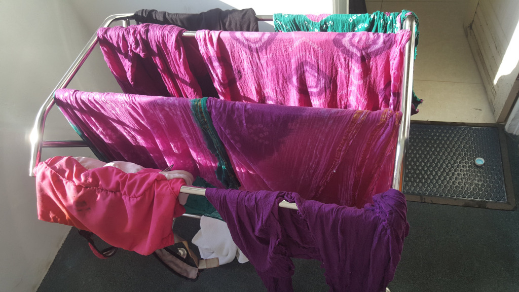 Clothes drying rack, positioned slightly off-centre, angled toward the lower right corner. The rack is loaded with a variety of garments, predominantly vibrant pink and purple textiles. These appear to be saris or similar long, flowing fabrics, some with tie-dye patterns exhibiting a soft, diffused coloration. The pinks range from a deep fuchsia to a lighter rose, while the purples are deep and rich. There's a smaller, folded garment of a coral-pink hue hanging on a lower section of the rack. Partially visible underneath and near the lower-left, are a darker pink item and a white piece of fabric (possibly a towel). A dark purple garment drapes predominantly over the lower right of the rack. The top level of the rack also shows a dark garment (likely black) slightly obscured by the vibrant pink fabrics underneath.