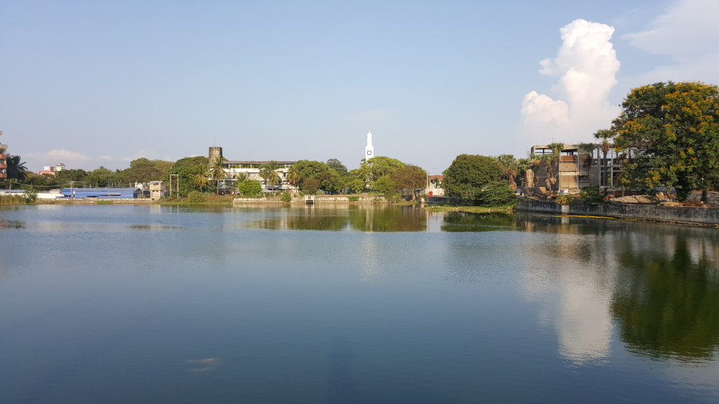 Calm, reflective body of water, possibly a lake or pond, occupying the majority of the foreground and middle ground. The water is a dark teal-green, its surface mirroring the sky and surrounding structures with remarkable clarity. The reflection of the clouds is particularly striking. The background features a low skyline of buildings, varying in height and architectural style. There’s a distinct white tower, resembling a clock tower or a slender minaret, that stands out against the greenery. Several buildings are low-slung, and one near the right bank appears to be under construction or in a state of disrepair, with exposed brick and scaffolding evident. The buildings are a muted mix of browns, creams, and grays.  Lush green trees and vegetation line the banks of the water, creating a natural border between the built environment and the aquatic space.