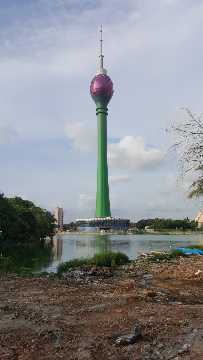 Central focus is the Colombo Lotus Tower, a tall, slender tower with a distinctive lotus-shaped top. The top is a rich, dark pink/purple, while the main body of the tower is a vibrant, Kelly green. It stands prominently against a partly cloudy sky. The tower is situated on a circular platform at the edge of a calm, greenish-brown body of water. In the foreground, a section of disturbed earth is visible, littered with debris and rubble, indicating ongoing construction or demolition. A few small fishing boats are visible on the water in front of the tower.