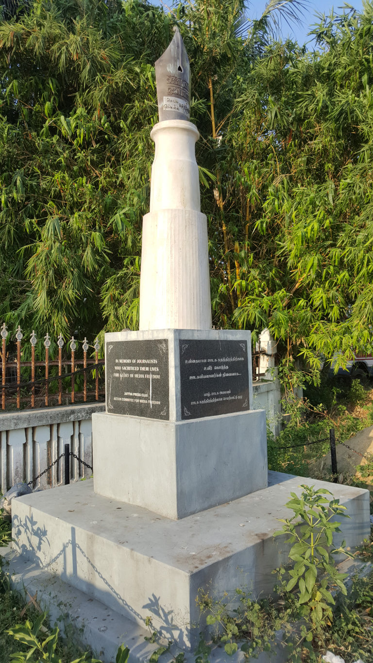 Tall, slender column of off-white, possibly marble or a similar material, resting on a square, light grey concrete base. The column is subtly fluted, and its top features a dark grey, metallic sculpture shaped like a fountain pen nib, angled slightly upward. Two dark rectangular plaques are affixed to the base of the column, one on each side, bearing inscriptions in both English and Tamil. A low, square platform of matching grey concrete surrounds the base, creating a raised area. A simple, black metal chain runs along the perimeter of this platform. Behind the monument is a dense wall of vibrant green bamboo, contrasting sharply with the grey tones of the structure.