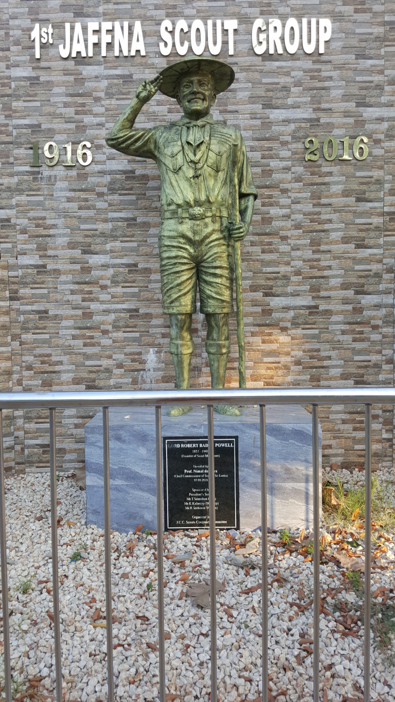 Bronze statue of Lord Robert Baden-Powell, founder of the Scouting movement. He is depicted in a Scouting uniform: a khaki short-sleeved shirt with badges, khaki shorts, and knee-high socks. He wears a wide-brimmed hat, slightly tilted, and his right hand is raised in a salute or gesture of greeting. He holds a staff or walking stick in his left hand. The statue stands on a rectangular, light grey stone pedestal.  Below the statue is a dark-grey or black plaque with engraved text detailing Baden-Powell's life and the unveiling of the statue. The statue is positioned against a wall of light-brown and beige textured stone tiles, arranged in a stacked, slightly irregular pattern, resembling a stone-clad building facade. Above and flanking the statue are metallic, gold-coloured numerals 1916 and 2016, suggesting a centennial commemoration. The words 1st JAFFNA SCOUT GROUP are prominently displayed above the statue in capital letters.