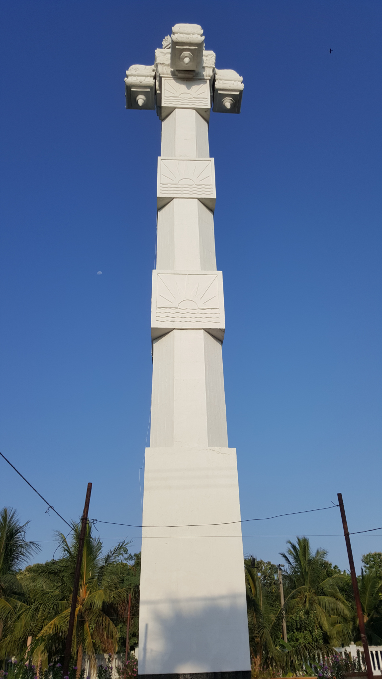 Tall, slender, white structure resembling a stylised obelisk or pillar. It's predominantly white with a series of recessed panels featuring a stylized sun and wave motif repeated at intervals along its height. The top of the structure is adorned with a more elaborate, cross-like or crown-like design, also white, with slightly darker shading suggesting shadow or texture. The structure stands against a vibrant, clear blue sky, almost devoid of clouds except for a tiny, distant crescent moon visible in the upper-left quadrant. The lighting suggests it's daytime, with the sun likely high in the sky, casting shadows that are barely visible on the pillar's surface. The background includes a sparse, tropical or subtropical landscape. Palm trees with long, slender fronds are visible at the bottom of the frame, suggesting a warm climate. There are also other less defined green trees and shrubs, and some thin, dark utility wires stretch across a portion of the lower part of the image. The overall colour palette is dominated by the intense blue of the sky, the stark white of the structure, and the various shades of green from the vegetation.