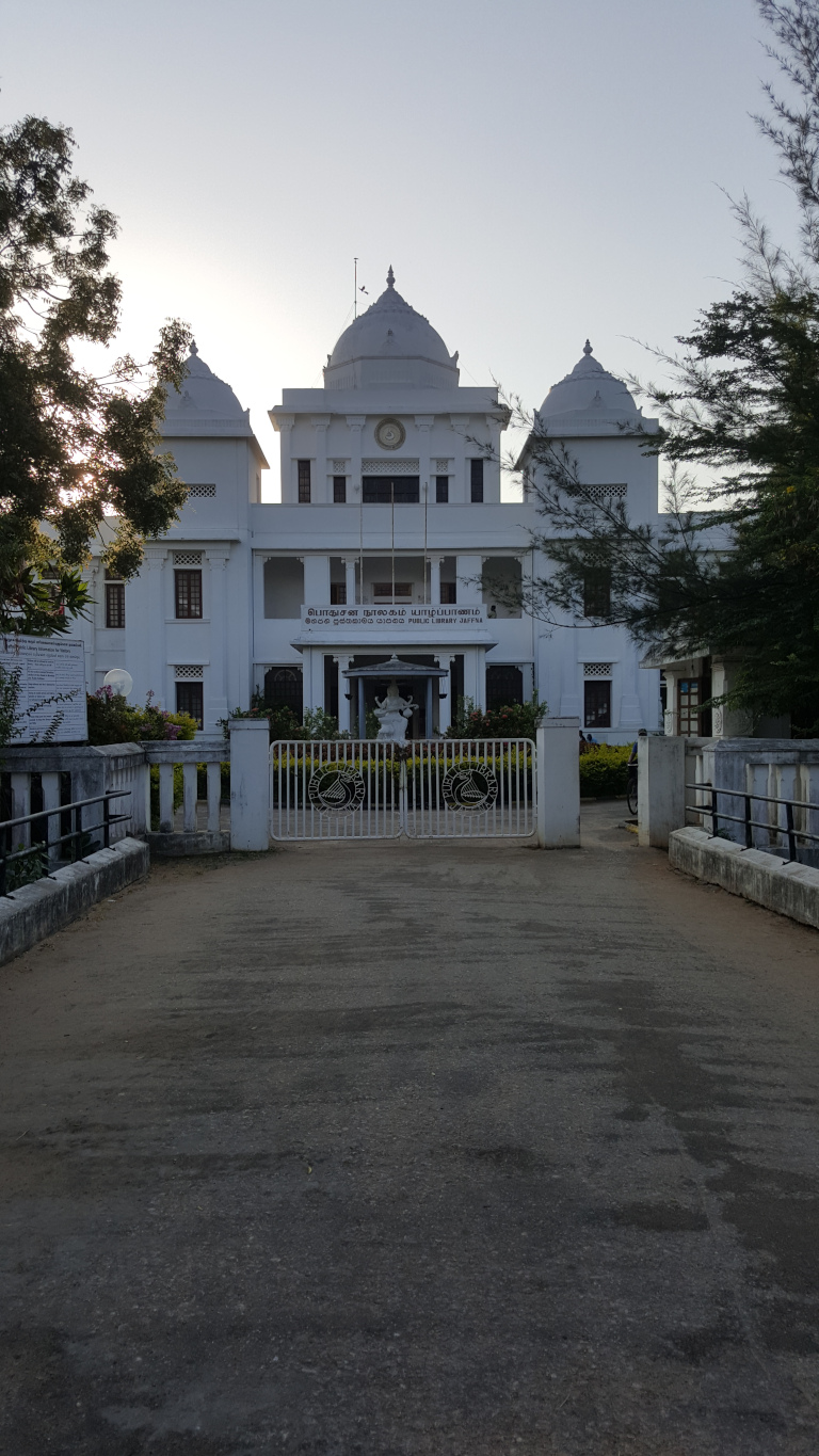 Grand, colonial-style white building, seemingly a public library, as indicated by signage in Tamil and English. It features symmetrical wings extending from a central section topped with a dome. Smaller domes crown the wings. A statue is visible within a small portico or recessed entryway in the central section. The building's entrance is guarded by ornate wrought-iron gates bearing a circular emblem. A few people are barely visible in the distance near the building’s right side. They appear small and indistinct, lacking detail, not interacting with the main features. The scene is set outdoors, with low-lying vegetation and trees surrounding the building. The trees are sparse, primarily seen on either side of the building and further back. The sky is a pale, almost washed-out light gray or pale blue, indicating either early morning or late afternoon light. The overall lighting is soft and diffused; there are no harsh shadows.