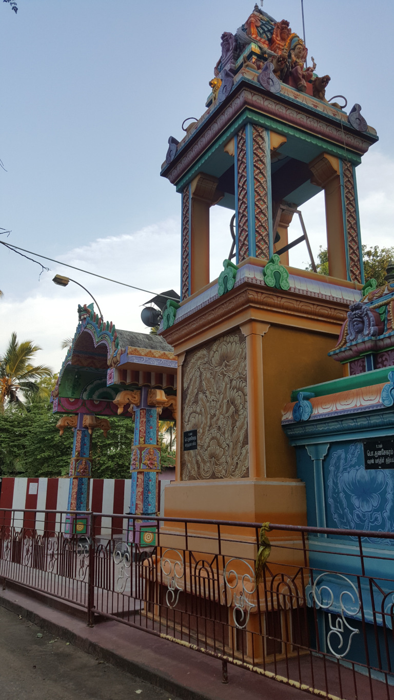 Multi-tiered, brightly coloured structure resembling a bell tower or shrine. It's predominantly orange and beige, adorned with elaborate carvings and painted murals featuring vibrant blues, greens, purples, and gold accents. Delicate, stylised floral and mythical creature motifs are woven into the carvings. Statues of deities or mythical figures are visible on the topmost tier, appearing in dynamic, slightly theatrical poses. A large bell is partially visible within the tower's open structure. To the left, a smaller, similarly styled gateway or entrance is visible, mirroring the architectural details of the main structure. Both structures are enclosed by a dark reddish-brown metal railing with ornate scrollwork incorporating stylised symbols along the base. A few sparse yellow-green strands of what look like decorative garlands or ribbons are draped on the railing.
