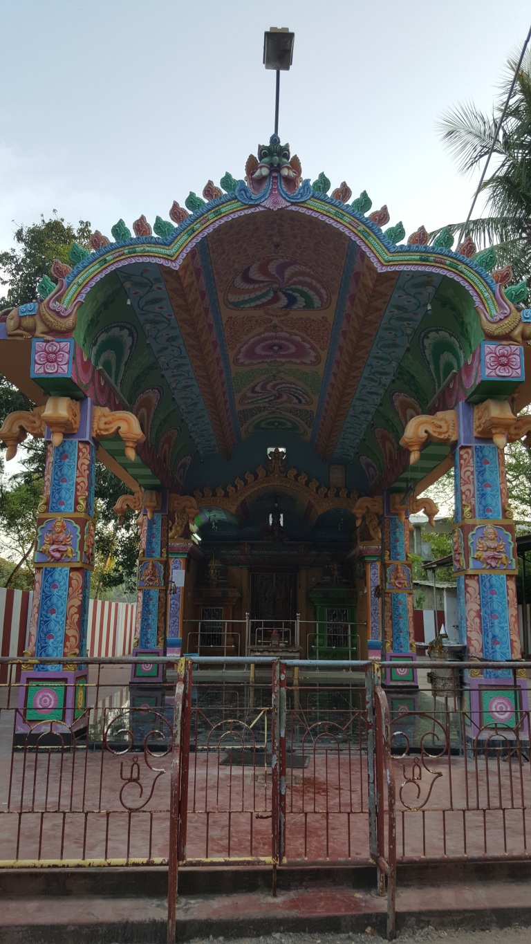 Vibrantly coloured Hindu temple structure, specifically what appears to be a mandapam (a covered pavilion) or entrance porch. he main focus is a colourful, intricately decorated structure with a curved, arched roof. The roof is painted in a swirling pattern of pastel greens, pinks, blues, and yellows, with decorative floral and leaf motifs. The pillars supporting the roof are similarly painted with detailed floral and mythical figure carvings. The colours are bold and slightly whimsical, almost cartoon-like in their bright saturation. In the foreground is a rusty, reddish-brown metal gate with a simple design, somewhat contrasting with the vivid temple structure behind it. The gate features a stylized Om symbol on each side. A glimpse of the temple's inner sanctum is visible through the gate. It appears to be more modestly decorated compared to the colourful entrance. No people are present in the image.