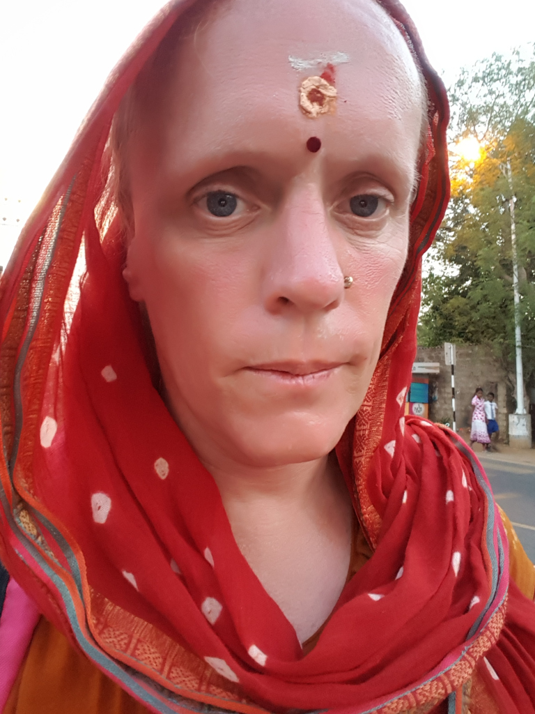 Close-up portrait of a fair-skinned woman, her face dominating the frame. She's wearing a vibrant red scarf or shawl, draped around her head and shoulders in a style suggestive of South Asian attire. The scarf is richly textured, featuring small, off-white dots scattered across its surface. A thin, multicoloured border runs along its edges. The woman appears to be directly facing the camera, her gaze is level and her expression is somewhat neutral, perhaps pensive or slightly melancholic. She has a bindi (a red dot) and a gold-colored marking on her forehead, common in Hindu traditions. A small, light-coloured piercing is visible in her left nostril. Her skin shows subtle redness, possibly from sun exposure.
