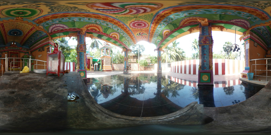 360 degree panoramic view of the interior and immediate exterior of a vibrantly painted Hindu temple. The main focus is a pavilion-like structure with a low, arched ceiling adorned with intricate, colourful frescoes. The pillars supporting the roof are richly decorated with similar vibrant designs. The floor is a highly polished, dark-colored stone, reflecting the surroundings. A small shrine, seemingly enclosed behind a railing, is visible within the pavilion's interior. Another shrine or structure is visible in the far background. The ceiling is a masterpiece of vibrant color, featuring swirling patterns, floral motifs, circular designs, and stylized depictions that evoke a sense of divine ornamentation. The colors include deep blues, greens, reds, yellows, oranges, and pinks, all intensely saturated. The columns are equally ornate, displaying vertical bands of detailed patterns and figures.