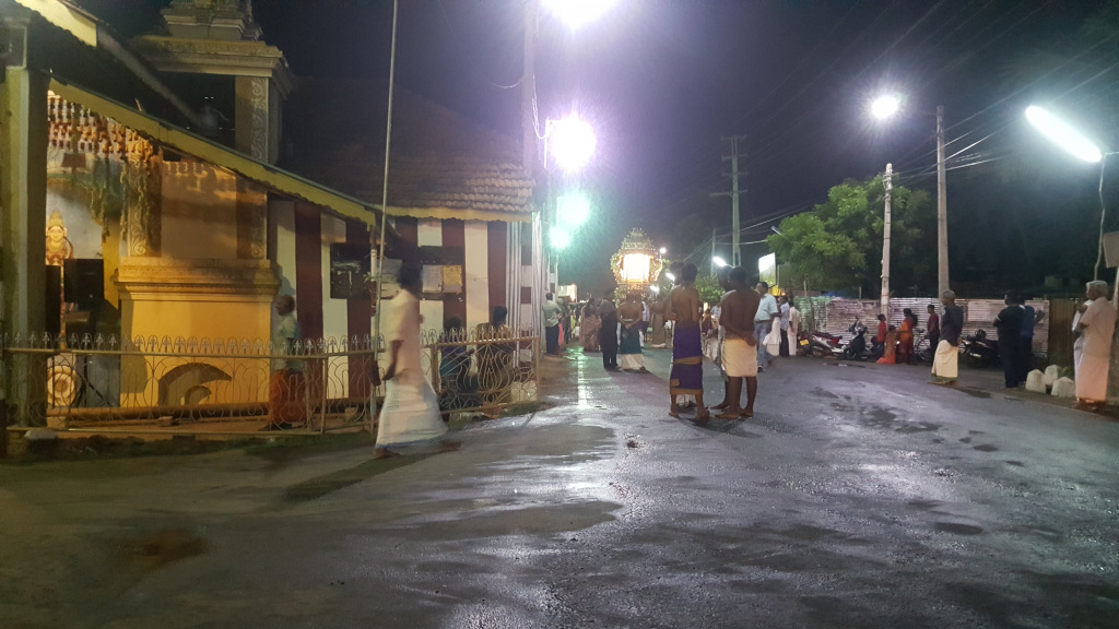 Street scene at night, seemingly outside a Hindu temple. A partially visible Hindu temple dominates the left side of the frame. Its architecture is ornate, with gold-toned details and carvings visible on the exterior walls and a tiered roof structure. A section appears to be an altar or shrine area, protected by an intricately designed wrought iron fence. Glowing, possibly decorative, elements suggest lighting from within. A group of approximately 20 people, mostly men, are positioned in the middle and right foreground. Several men are clad in white dhotis (traditional South Asian garments), seemingly participating in a religious procession or ritual. Their postures suggest they are either observing or pausing. Some appear bare-chested, and others are dressed in more casual attire. Women are sparsely visible, often partially obscured in the background, dressed in darker, less-distinct clothing. One group of people are gathered near motorbikes, which are parked to the right. A slightly blurry structure, suggesting a palanquin or float typical of religious processions, is visible in the mid-ground. This indicates a festival or event may be taking place.