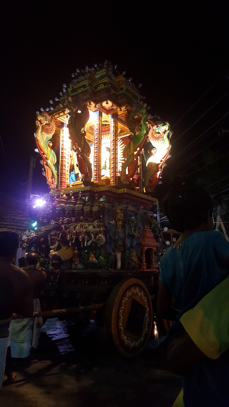 Brightly lit, ornate temple chariot (rath). It's a multi-tiered structure, seemingly made of wood and richly decorated with vibrant colours—greens, reds, golds, and blues—intricate carvings depict mythical figures and animals, and small lights are embedded along its upper edges, creating a glowing halo effect. The chariot is on wheels, one of which is prominently featured in the foreground. The wheels are large, wooden, and painted with geometric patterns in dark brown, orange, and gold. Several people are visible, though their faces are largely obscured by shadows. Two men, seemingly bare-chested or wearing minimal clothing (one in a white dhoti), are positioned near the front of the chariot, appearing to pull or guide it.  A child or young person, with dark hair, is slightly behind and to the right, gazing at the chariot with a seemingly contemplative expression.  Their clothing is a teal blue top. Another person, partially visible, is behind the child, wearing a yellowish garment. Their actions suggest participation in a religious procession or festival.