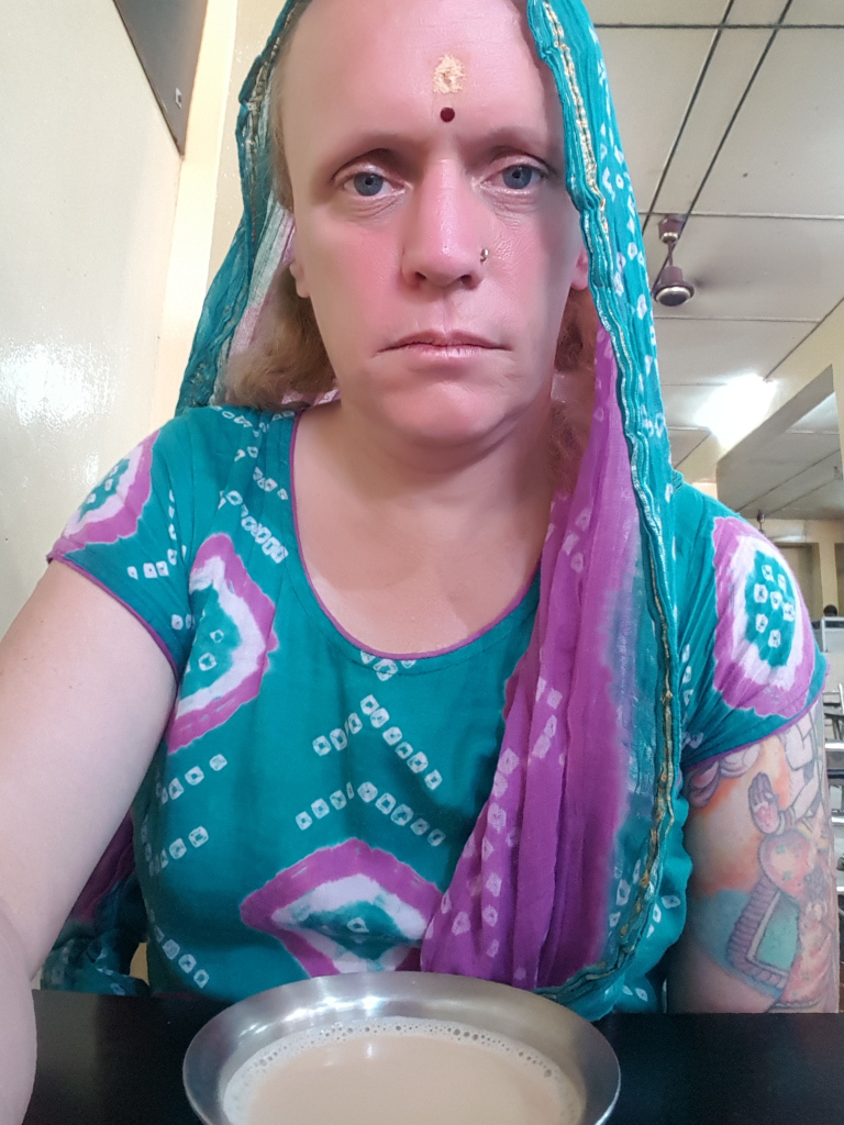 Fair-skinned woman, possibly in her 30s or 40s, with a calm, almost pensive expression. She's draped in a vibrant teal and purple dupatta (a long scarf), which is loosely arranged around her shoulders and neck. The dupatta features a paisley-like pattern with a subtle sheen. She wears a teal kurta (a long shirt) with a similar, but less pronounced, paisley pattern in shades of purple and white. Her arms are visible, with a noticeable tattoo on her right upper arm, appearing to be a stylized cartoonish or illustrative design.  She has a small gold bindi (a decorative dot) on her forehead and a small nose piercing. The woman is seated, appearing to be in a cafe or eatery, with a metallic, shallow bowl in front of her containing a light brown liquid (likely tea or coffee). Her gaze is directed slightly downward, and her posture is relaxed but still. She is not actively doing anything beyond seemingly observing her surroundings.