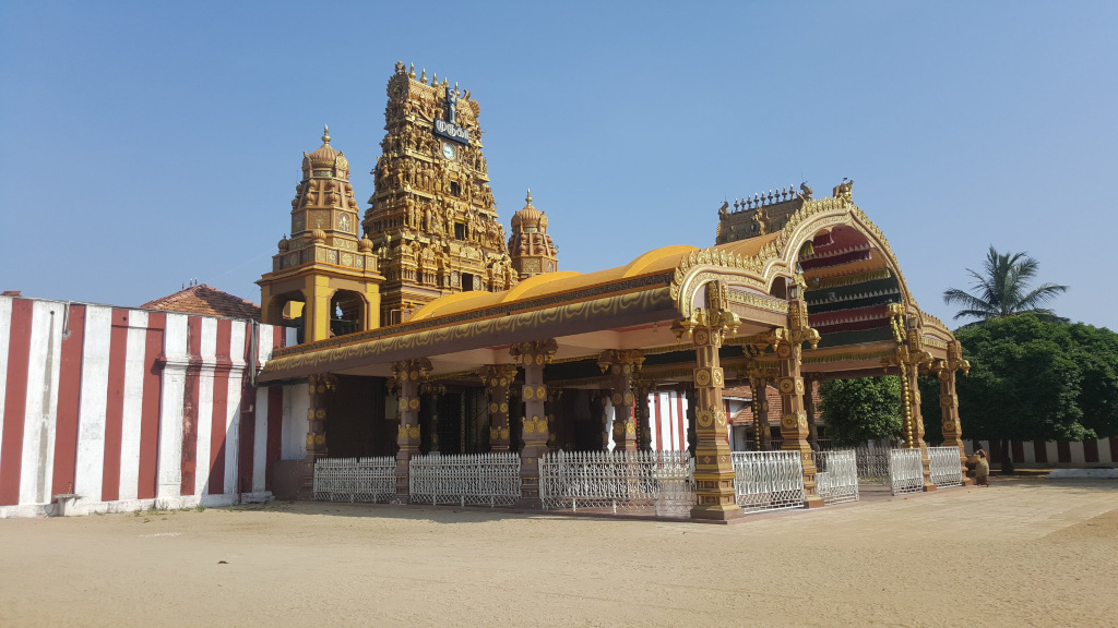 Resplendent Hindu temple, predominantly gold and brown, exhibiting intricate carvings and ornamentation. Its gopuram (tower) rises majestically, multi-tiered and adorned with countless miniature sculptures and decorative elements. The gopuram's gold paint shimmers under the light, reflecting a warm golden hue.  Adjacent to the main structure is a large, ornate, covered pavilion, also exhibiting golden coloring and elaborate carvings. This pavilion sits on sturdy, ornately carved pillars, creating a grand entrance or waiting area. A simpler, red and white striped wall forms a partial backdrop to the left. A single, indistinct figure, seemingly a person, is barely visible near the right bottom corner of the temple complex, engaged in an action too small to discern. The scene is bathed in the bright, clear light of a sunny day. The sky is a brilliant, unblemished blue, contributing to the overall feeling of clarity and warmth. The temple complex stands on a pale tan, sandy ground that extends to the foreground. In the background, beyond the temple's immediate surroundings, a lush green tree partially obscures what appears to be a further building. The dominant colors are the intense gold of the temple's structure and the deep red and crisp white of the adjacent wall, providing a powerful contrast against the muted tones of the sandy ground and the green foliage.