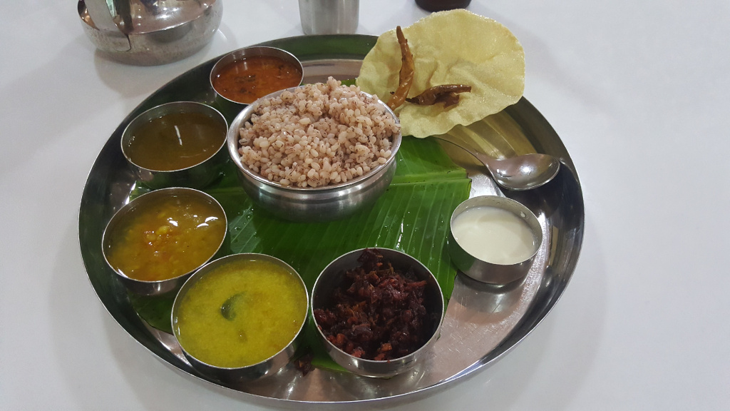 Large, circular, stainless steel thali (tray) brimming with a South Indian meal. Nestled on a vibrant green banana leaf within the thali is a bowl of light-brown, slightly speckled rice or grain, possibly barnyard millet. Surrounding the rice are six small, similarly styled stainless steel bowls. These contain various curries and accompaniments.