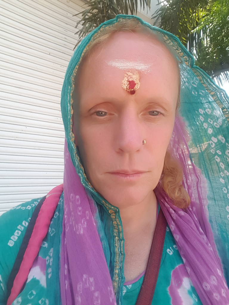 Close-up selfie of a fair-skinned woman with blonde hair partially visible beneath a vibrant teal and lavender dupatta (a type of scarf). The dupatta is draped over her head and shoulders, partially obscuring her hair. She appears to be standing still, looking directly at the camera with a neutral, almost pensive expression. Her forehead displays a red and gold bindi (a decorative mark worn by Hindu women), suggesting cultural or religious significance. A small, gold-colored nose stud is also visible. She's wearing a teal and white tie-dye style top, and part of a maroon shoulder bag strap can be seen.