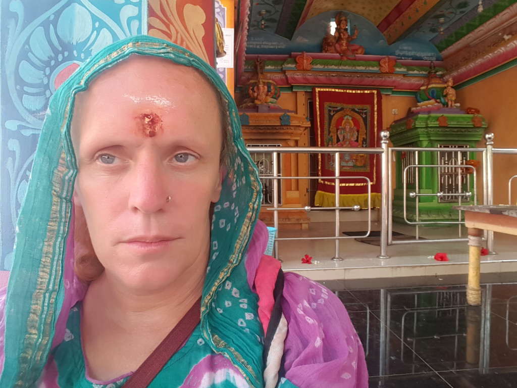 Fair-skinned woman, appearing to be middle-aged, with a contemplative expression. She's wearing a vibrant teal and purple shawl or dupatta draped over her head and shoulders, partially obscuring her hair. The shawl features intricate, repeating patterns in a lighter teal. Beneath the shawl, a purple and teal patterned top is visible. She has a small, dark mole or piercing on the left side of her nose. Most notably, she has a reddish-brown, slightly raised mark on her forehead, suggesting a bindi or a recent injury. She appears to be still and gazing slightly off to her left, directly at the viewer. She carries a brown strap across her torso. The woman is positioned in the foreground, sharply in focus, against a brightly coloured background. The background depicts the interior of what seems to be a Hindu temple or shrine. The walls are adorned with vivid murals featuring deities and intricate, ornate patterns in warm oranges, yellows, greens, blues, and reds. Metallic silver railings run horizontally across much of the background, separating sections of the temple's interior. Small shrines with colourful statues are visible in the background. The floor is dark, polished tile, reflecting some of the scene's bright colors. The lighting appears to be natural daylight, streaming into the temple space, casting no harsh shadows on the woman. The overall color palette is bold, warm, and rich, with a strong contrast between the woman's attire and the temple's decoration. A few scattered red flowers are visible on the temple floor.