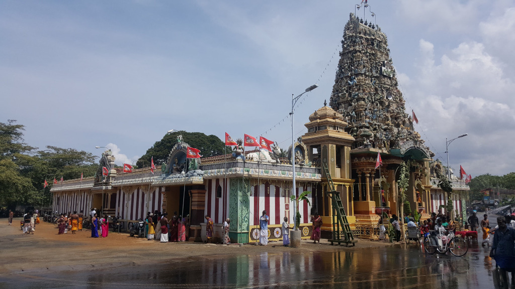 Vibrant Hindu temple, a complex structure dominated by a tall, ornate gopuram (tower). The gopuram is densely decorated with colourful sculptures and carvings of deities and mythical figures, creating a multi-layered, almost chaotic visual texture. Its colors are a mix of warm yellows, oranges, and greens, contrasting with the grey stone. Red flags with white symbols flutter from various points on the temple. The temple complex also includes a long, lower building with a striped red and white facade, suggesting different sections within the temple's grounds. Many people, mostly dressed in brightly coloured South Asian clothing, are gathered around the temple, some appear to be worshipping or participating in a religious event.  They are standing in small groups, near entrances, or walking along the wet ground. A few rickshaws or cycle-rickshaws are parked near the temple. A ladder leans against a portion of the temple structure, suggesting ongoing maintenance or preparations.
