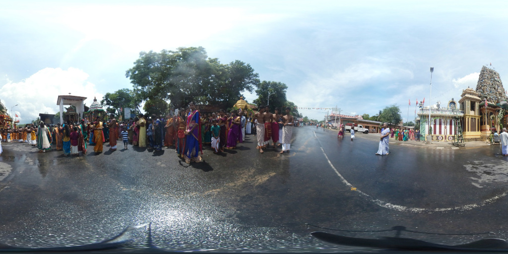 360-degree panoramic view, centred on a procession of people on a wet road. The procession is composed of a multitude of individuals predominantly dressed in vibrant South Asian attire, a mix of sarees in various colours (reds, pinks, oranges, blues, greens), and men wearing dhotis and kurtas, many bare-chested. Several women wear richly coloured sarees with elaborate borders and adornments. The group is dense in the centre of the frame, tapering off towards the edges. They appear to be participating in a religious festival or ceremony. The main focus is on the women and men in the central area, with their movement conveying a sense of procession or pilgrimage. Towards the far right, several individuals stand near a temple, and near the left, another group is positioned seemingly awaiting its turn in the line.A significant object is a partially visible ornate palanquin or chariot, which suggests a religious significance to the event. Several men, bare-chested and appearing to be carrying or supporting this chariot, occupy the central area amongst the women. The procession is flanked by buildings on either side, with a larger temple structure more prominently visible on the right.