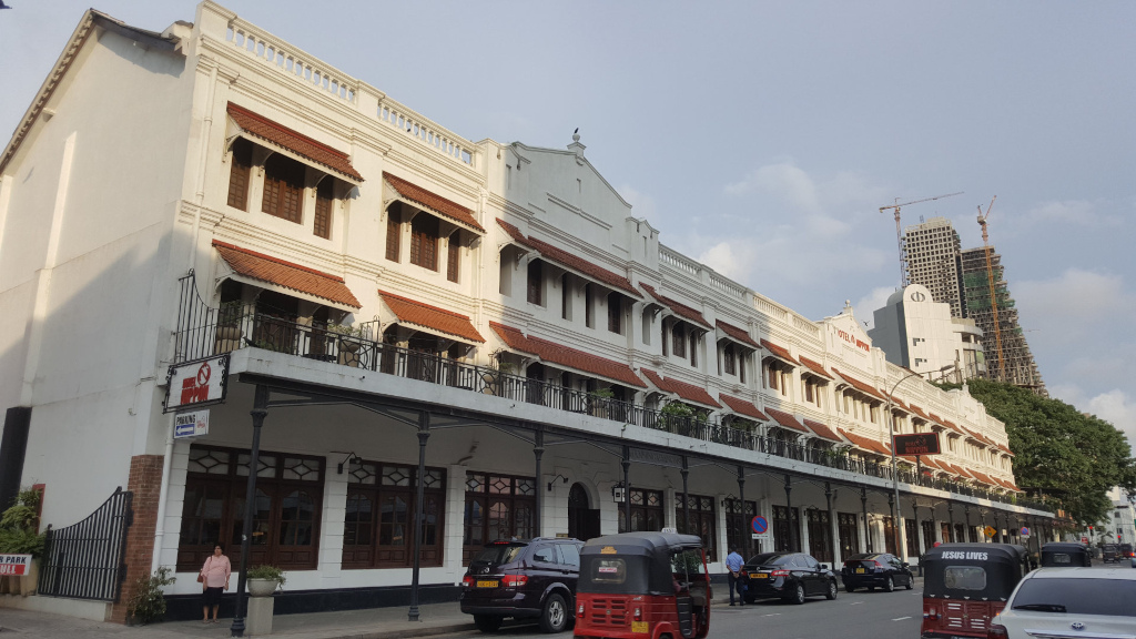 Long, low, colonial-style building, creamy white with terracotta-tiled awnings over the windows and balconies. The building stretches almost the entire width of the frame, receding into perspective. It displays a uniform architectural style, suggesting a hotel or a series of connected shops. A dark, wide metal awning extends along the entire length of the building’s ground floor, creating a covered walkway.