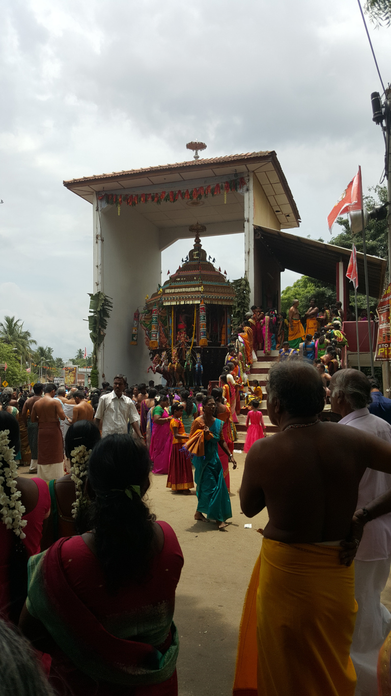 Large, ornate chariot-like structure, a Ther, typical of South Indian temple festivals. It's intricately decorated with vibrant colours—reds, golds, greens, and blues—and features miniature figures and embellishments. The Ther is positioned under a simple, white-walled, roofed structure that seems to be a temporary setup for the procession. A large crowd of people surrounds the Ther. Many are dressed in brightly colored traditional South Indian attire—saris and dhotis in a variety of hues and patterns. Some women are adorned with jasmine flower garlands in their hair. The people's actions suggest participation in a religious festival: some stand observing; others seem to be involved in the preparations or procession of the Ther. A significant number of men are shirtless, or wear only a dhoti, a common sight in South Indian religious events. One man in the foreground prominently displays his bare back, wearing only a bright yellow dhoti, and slightly turned towards the Ther.