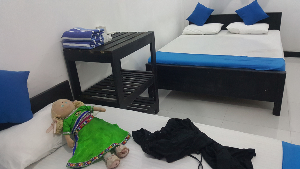 Small, rag doll dressed in a vibrant green and multicoloured outfit, reminiscent of traditional South Asian clothing. She’s lying on a white bed with her head resting on a white pillow.  Her pose is relaxed, limbs slightly askew. Near the doll, a black garment, possibly a dress or top, lies crumpled on the bed. A stack of blue and white striped towels sits neatly on a dark brown wooden side table. Two blue square throw pillows rest on a black double bed in the background, implying it's unmade, though linens are present.  Another blue pillow sits on the bed in the foreground, against the white wall. The bed is simple and boxy in its design. The side table is small and rectangular, with open shelving.