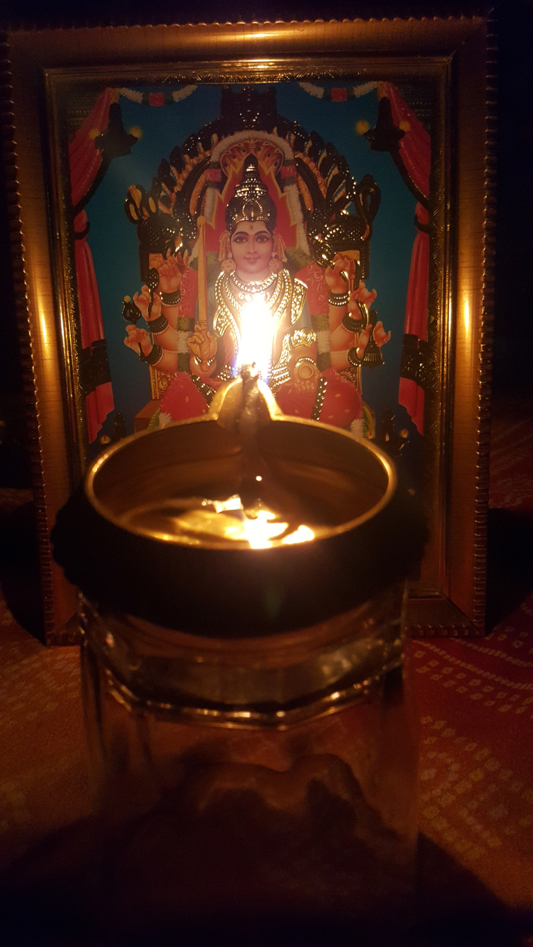 Small, brass oil lamp (diya), glowing warmly. Its circular bowl, filled with flickering oil and a bright flame, sits atop a clear, cylindrical glass container. The lamp is positioned directly in front of a framed picture. The picture depicts a Hindu deity, possibly a female goddess, seated regally on an ornate throne. The goddess is richly adorned with gold jewelry and vibrant clothing, rendered in a style reminiscent of traditional Indian religious art. The colours are bold – reds, golds, blues, and a touch of green in the background. 