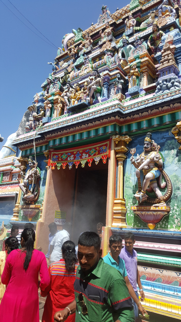 Vibrantly coloured Hindu temple, its entrance partially obscured by a gathering of people. The temple's gopuram (tower) is heavily decorated with numerous intricately carved and painted figurines of deities and mythical creatures. These figures are predominantly in shades of blue, green, gold, red, and white, creating a striking polychromatic effect. The architecture displays a multi-tiered structure with elaborate ornamentation. A partially visible sign in a local language is affixed to a pillar near the entrance.