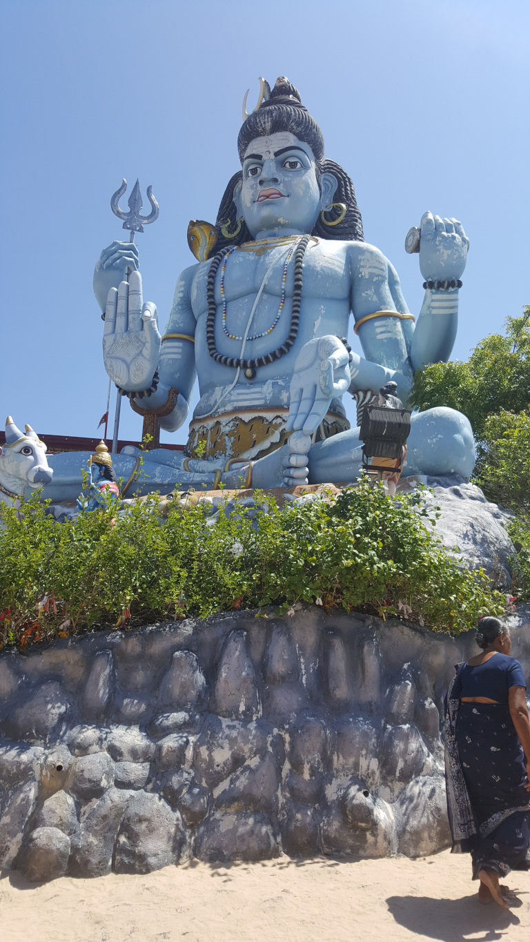 large, imposing statue of Shiva, the Hindu god. The statue is light blue, almost greyish-blue, with darker accents defining its muscular physique and facial features. Shiva is depicted seated in a meditative pose, holding a trishul (trident) in his upper right hand and displaying a stylised hand gesture (mudra) with his left. He wears a beaded necklace and ornate armbands. His expression is serious and slightly stern. At the base of the statue, partially obscured by vegetation, a smaller Nandi (bull) statue is visible. To the lower right, a woman is standing. She's dressed in a dark blue, almost black, sari with a patterned design. Her skin tone is dark brown. She appears to be observing the Shiva statue, her posture relaxed but attentive.