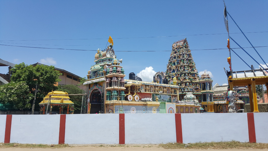 Vibrant Hindu temple complex, seemingly under some construction or renovation.  Two distinct gopurams (tower structures) dominate the scene. The closer one is a multi-tiered structure, richly adorned with colourful, intricate carvings and statues of deities and mythical figures. Its colours are a bright mix of yellows, blues, greens, reds, and whites, giving a sense of festivity. This gopuram has a relatively small, open gateway visible in the centre. The second, taller gopuram, set further back, is similarly decorated but shows a more weathered, perhaps older, look. In the foreground, a low white wall with vertical red stripes separates the viewer from the temple. A small, golden, ornate chariot-like structure is partially visible behind the wall, to the left of the main gopuram. To the right of the main temple structure, another smaller pavilion-like structure with a wooden roof is also visible. Thin electrical wires run across the sky above the temple. There are no people clearly visible in this image.