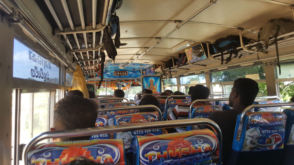Interior shot of a crowded Sri Lankan bus. The bus is filled with people, predominantly men and women with dark hair, seated closely together. Most are looking forward, appearing to be on a journey. Their postures are relaxed, some leaning slightly, indicative of a long ride. Their clothing is casual and varies. One person's backpack is visible on the overhead rack. The seat backs prominently display vibrant advertisements for a travel company named Thusha Travels, featuring colourful graphics and contact information.