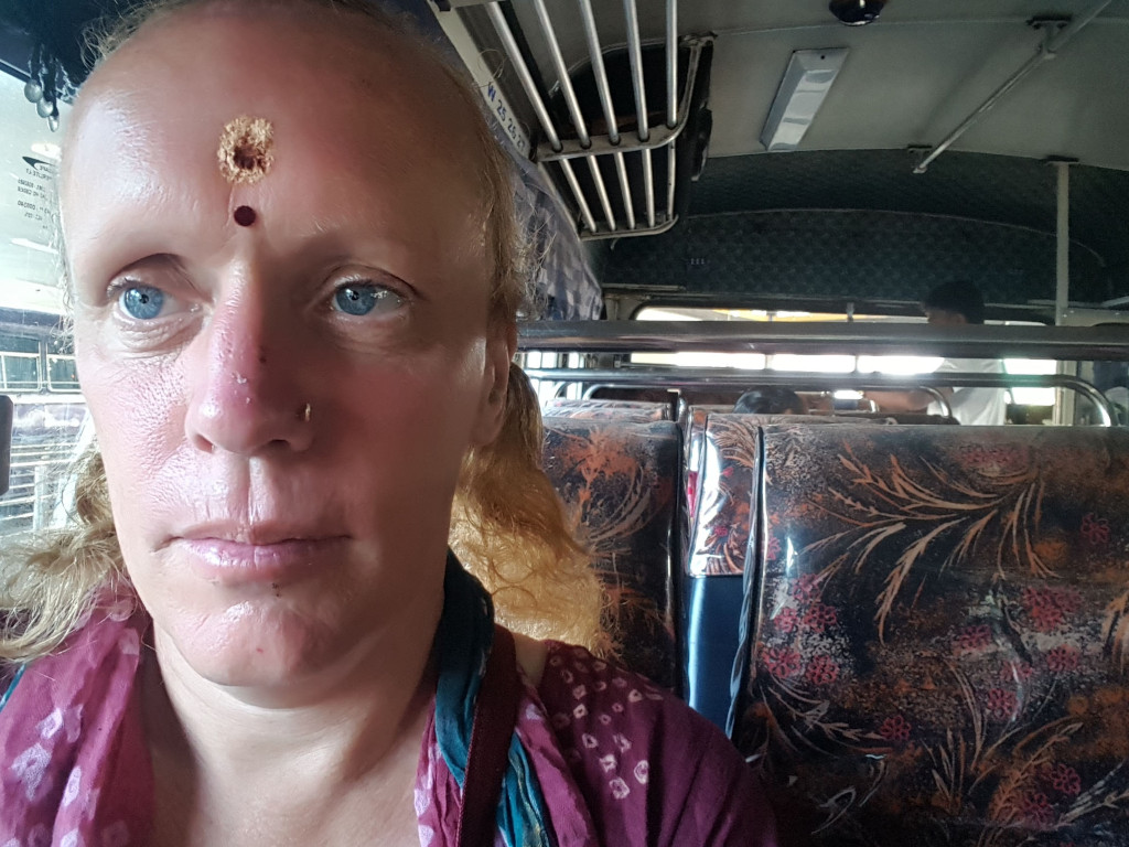 Close-up selfie of a fair-skinned woman with shoulder-length, slightly wavy light-brown hair. She appears to be in her 40s or 50s. Her face is the central focus, taking up most of the frame. She has blue eyes, a prominent nose with a small nose ring, and a noticeable round, light-brown mark or wound on her forehead, just above the centre. A small red bindi is placed just below this mark. Her expression is serious, almost sombre; her lips are slightly pursed. She's wearing a maroon-coloured garment with a teal scarf or shawl draped around her neck and shoulders. The woman is sitting in a bus, slightly turned to the left, and her gaze is slightly downward and toward the viewer. In the background are blurry figures and the interior of a bus.